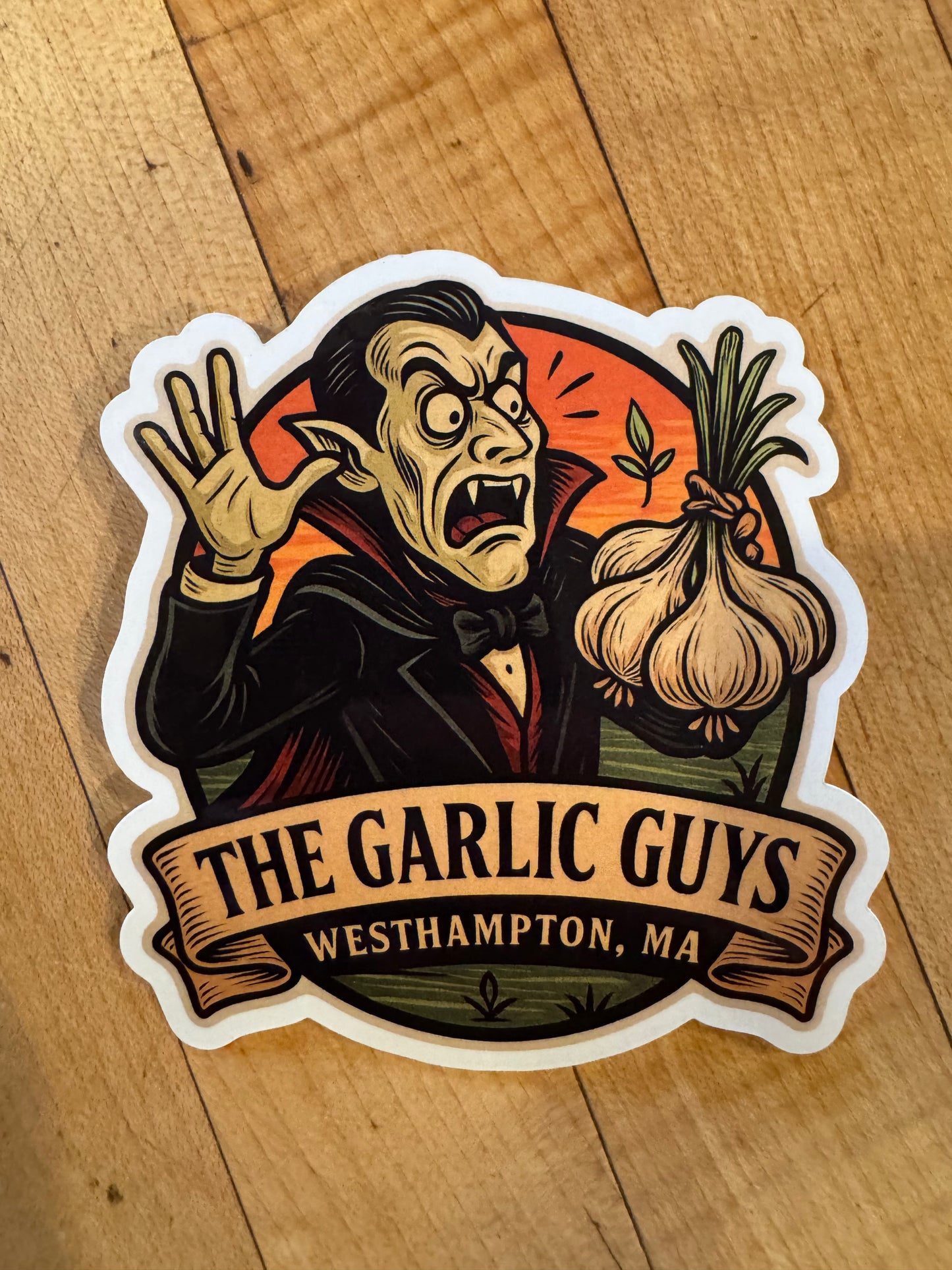 Sticker of a vampire holding garlic with 'The Garlic Guys' text on a wooden surface
